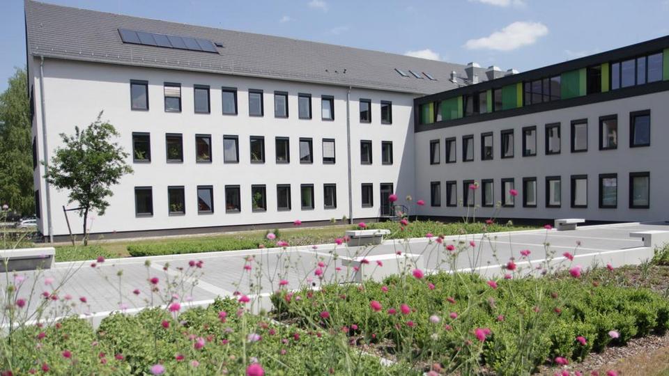 Modernes weißes Bürogebäude mit rechteckigen Fenstern und einem flachen, begrünten Innenhof im Vordergrund. Im Vordergrund blühen pinkfarbene Blumen, dahinter sind gepflasterte Wege und niedrige Beete zu sehen. Das Gebäude besteht aus mehreren, miteinander verbundenen Trakten. Der hintere Trakt hat ein Satteldach, der angrenzende Trakt ein Flachdach. Der Himmel ist blau mit wenigen Wolken.