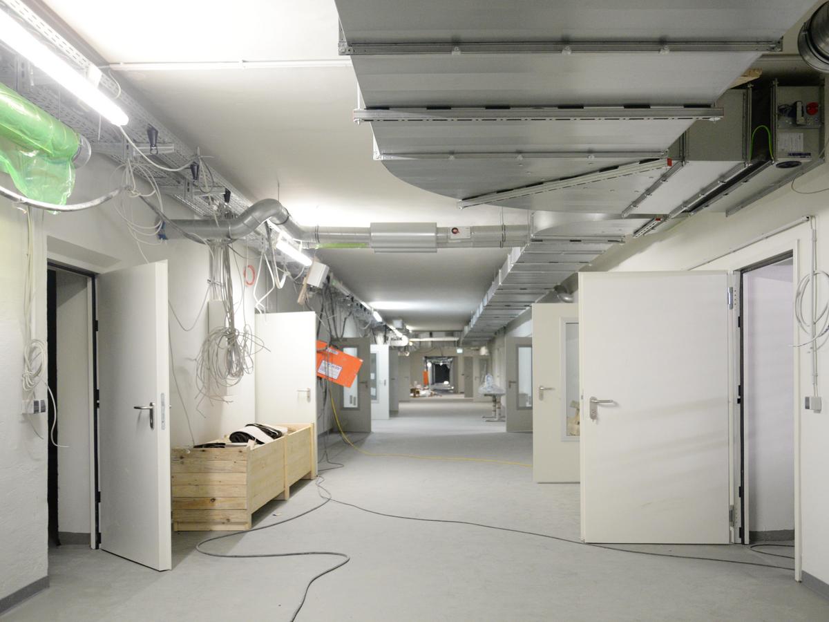 Blick in einen langen Flur mit vielen geöffneten Türen rechts und links. An der Decke viel Technik, Lüftungsleitungen, etc. Es ist Baustelle mit offenen Kabeln und etwas Unordnung.