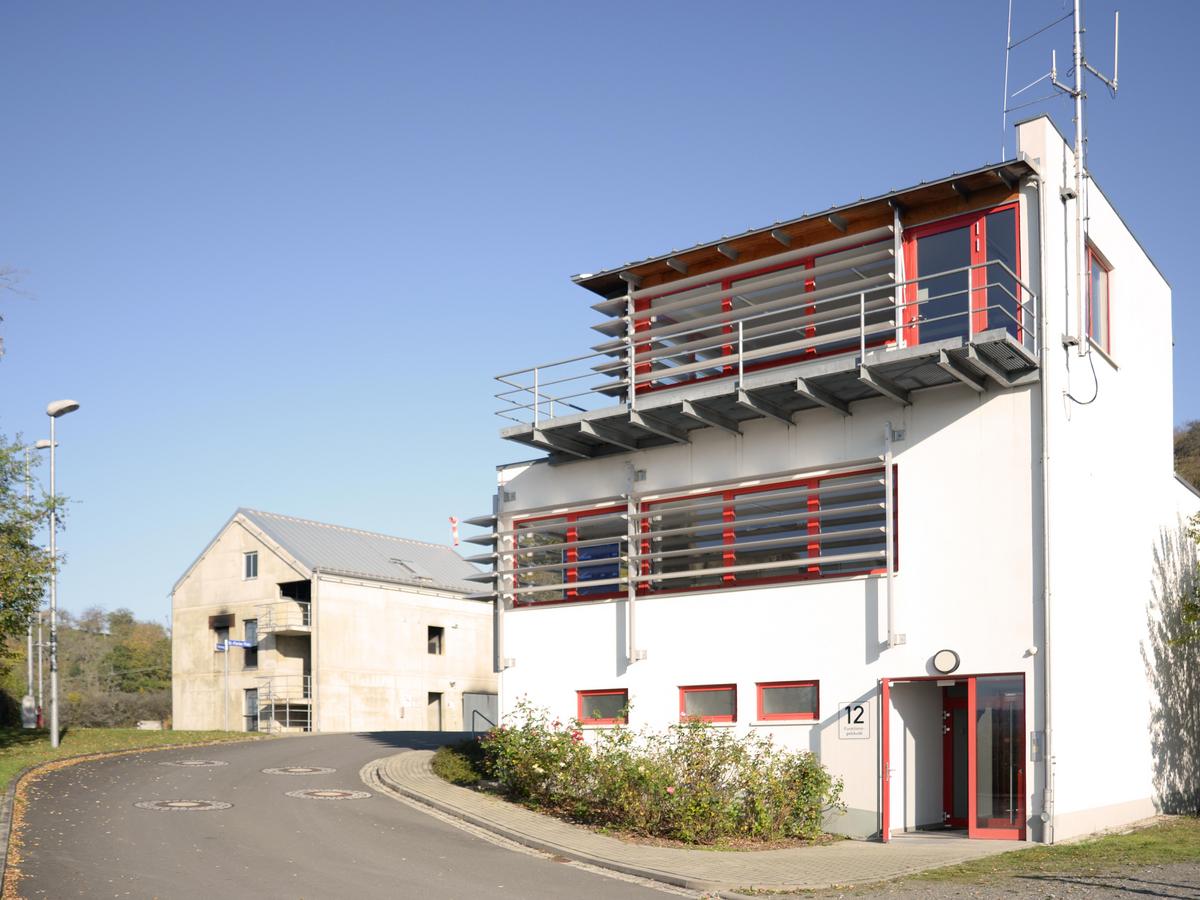 Eine Straße führt an zwei Gebäuden vorbei. Rechts im Vordergrund ein weißes dreigeschossiges Gebäude mit roten Fensterrahmen und horizontalen Sonnenschutzlamellen. Im Hintergrund der Betonbau zur Übung mit Rußflecken an der Fassade.