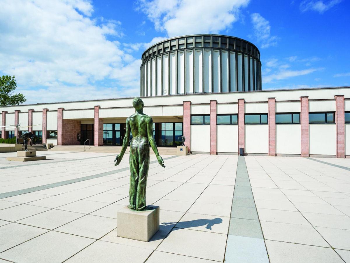 Foto vom Vorplatz, eine Kupfer-Statue auf profanem Sockel im Vordergrund. Die Figur öffnet sich. Über den Platz blickt man auf den flachen Eingangsbau mit seiner vertikalen Gliederung aus gemauerten Stützen aus roten Sandstein. Dazwischen läuft ein Fensterband. Im Hintergrund der Rundbau des Panorama Museum, wie ein gigantisches Zahnrad in weiß. Blauer Himmel mit Wolken.