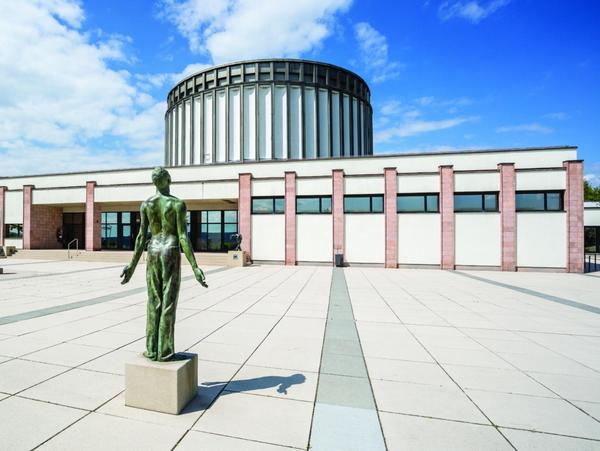 Foto vom Vorplatz, eine Kupfer-Statue auf profanem Sockel im Vordergrund. Die Figur öffnet sich. Über den Platz blickt man auf den flachen Eingangsbau mit seiner vertikalen Gliederung aus gemauerten Stützen aus roten Sandstein. Dazwischen läuft ein Fensterband. Im Hintergrund der Rundbau des Panorama Museum, wie ein gigantisches Zahnrad in weiß. Blauer Himmel mit Wolken.