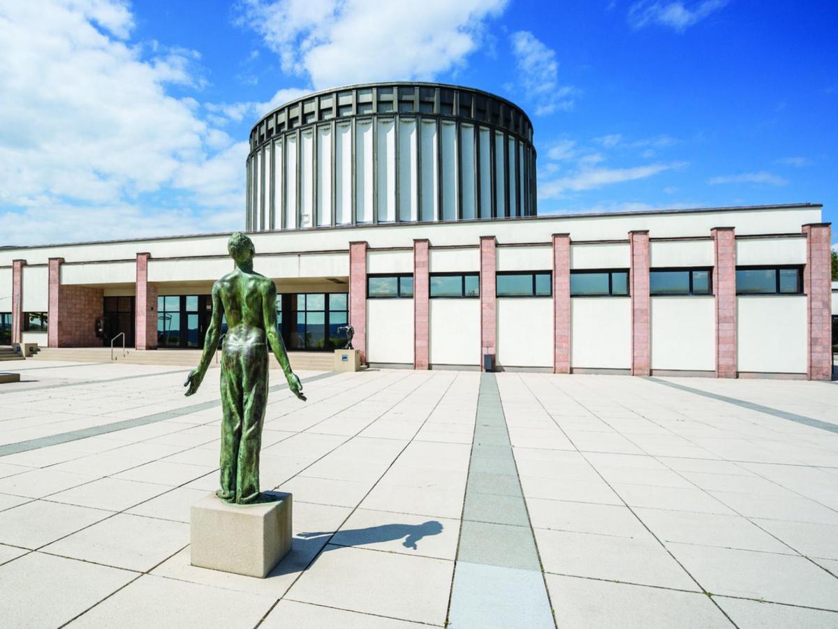 Foto vom Vorplatz, eine Kupfer-Statue auf profanem Sockel im Vordergrund. Die Figur öffnet sich. Über den Platz blickt man auf den flachen Eingangsbau mit seiner vertikalen Gliederung aus gemauerten Stützen aus roten Sandstein. Dazwischen läuft ein Fensterband. Im Hintergrund der Rundbau des Panorama Museum, wie ein gigantisches Zahnrad in weiß. Blauer Himmel mit Wolken.