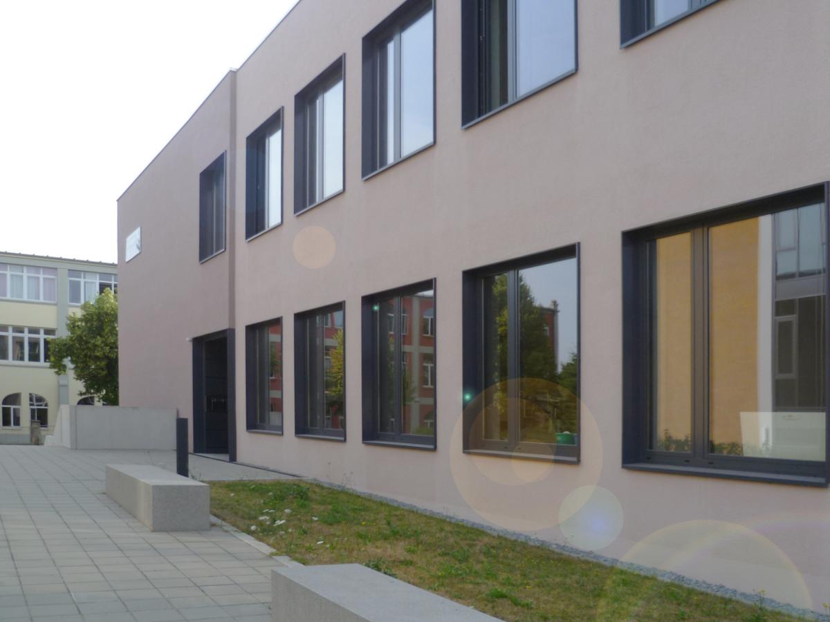 Foto von der Zuwegung zum Gebäude. Betonsitzbänke stehen an der Grünfläche, die dem Gebäude vorgelagert ist. Die Fenster im Raster dominieren das Foto. Es ist heiter, keine Personen zu sehen.