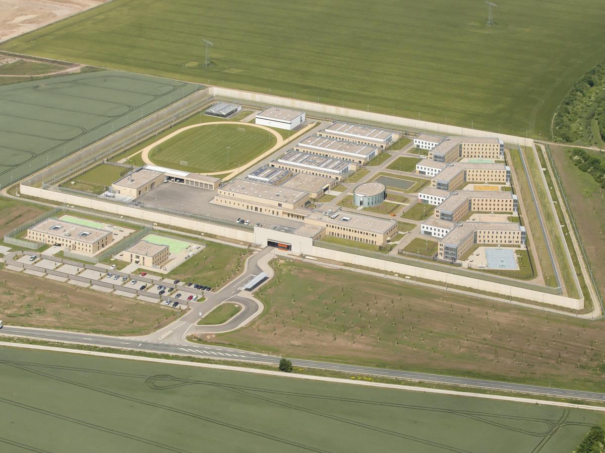 Luftbild der trapezförmigen Anlage, rundherum Grünfläche ohne Details erkennen zu können. Man erkennt deutlich den Sportplatz in der Anlage und die Staffelung der Gebäude an der schrägen Trapezseite. Unterhalb verläuft von links nach rechts eine Straße. Zwei Gebäude liegen außerhalb der Anlage mit einigen Parkplätzen.