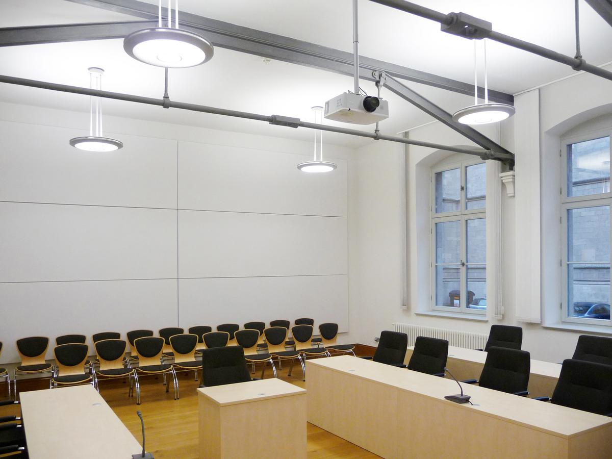 Kleiner Verhandlungsraum mit Stahlkonstruktion, sichtbar, grau lasiert, unterhalb der weißen Decke. Rückwand mit Schallschutzpanelen weiß. Runde Leuchten hängen von der Decke herab. An der Rückwand zwei Reihen mit schwarzen Stühlen für Zuhörer. Im Vordergrund die Holzpulte, als Kuben für die Gegnerparteien.