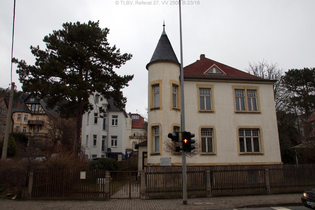 Straßenansicht mit Ampel im Vordergrund, eingezäuntes Grundstück mit zweigeschossigem Gebäude mit Turmanbau