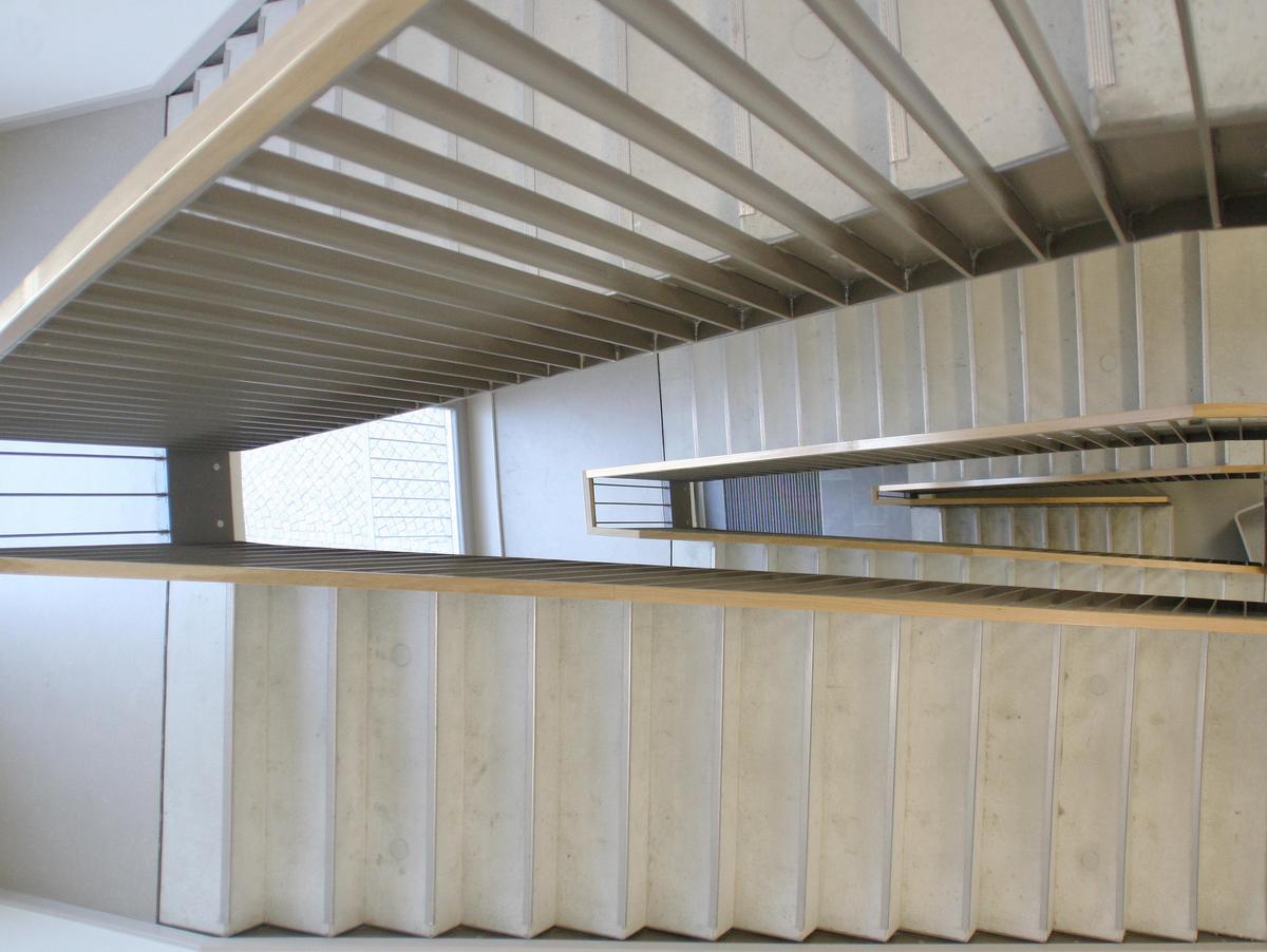 Foto vom Treppenpodest nach unten in das Treppenauge über drei Geschosse. Heller Bodenbelag, Handlauf eckig aus Holz und Geländer aus vertikalen Flachstahl in anthrazit.