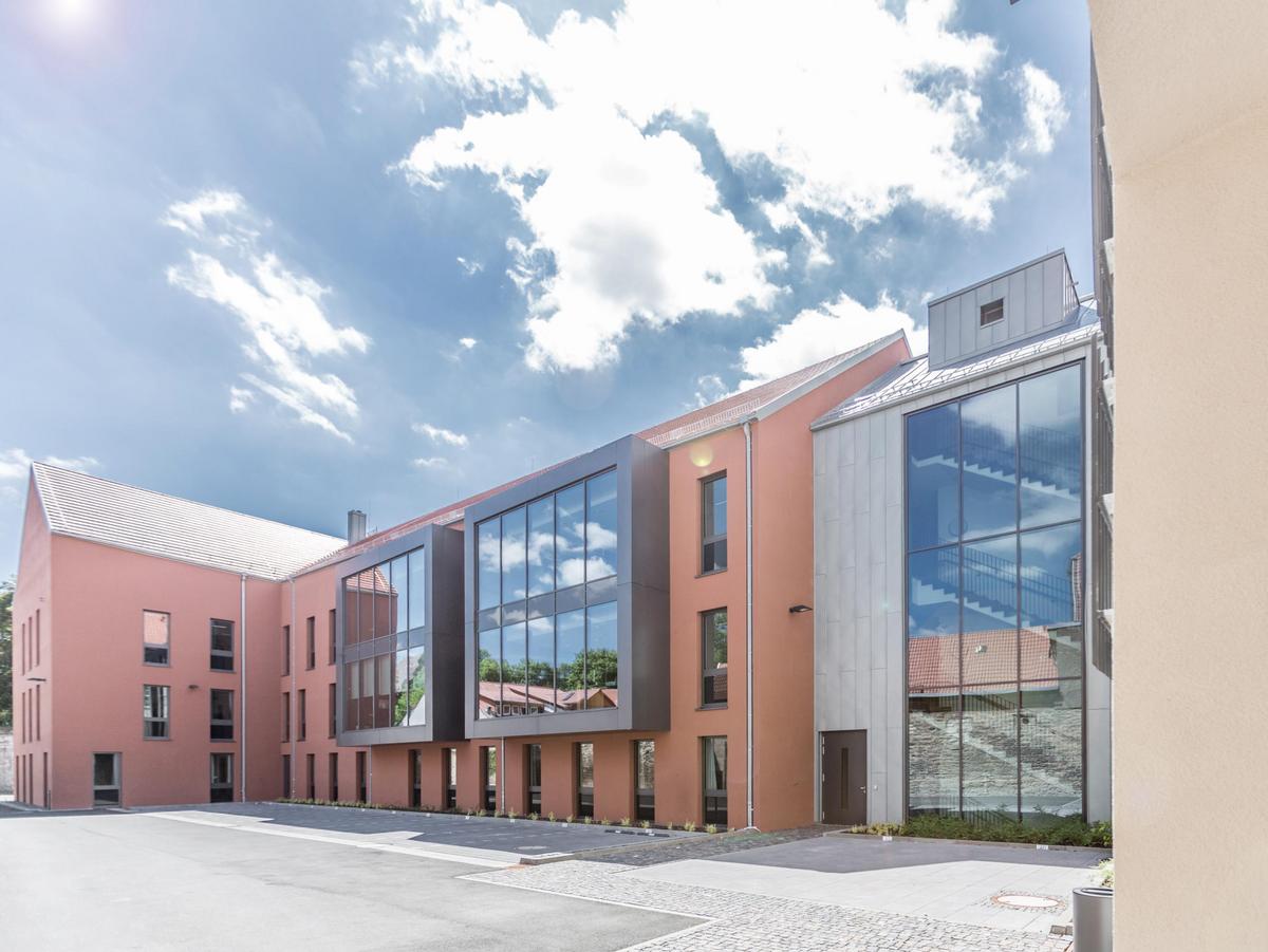 Dreigeschossiger orangener Neubau mit Satteldach. Die Fensterelement sind rasterförmig angeordnet und wirken schwarz. Zwei moderne quadratische graue Erker schieben sich aus der Fassade. Die Dachfläche ist grauen Dachziegeln belegt. Im Vordergrund eine befestigte Fläche und Parkplätze, rechts der Verbindungsbau aus Titanzink und viel Glas. Heiteres Wetter. Wie Bild 1 jedoch sieht man rechts am Bildrand noch teile des Altbaus in hellorange.