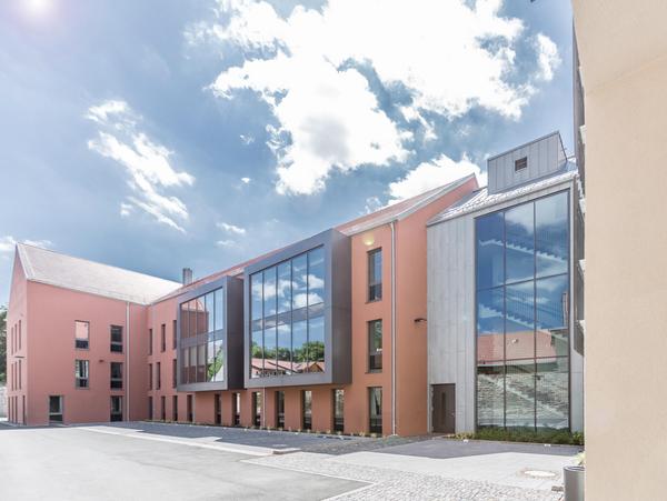 Dreigeschossiger orangener Neubau mit Satteldach. Die Fensterelement sind rasterförmig angeordnet und wirken schwarz. Zwei moderne quadratische graue Erker schieben sich aus der Fassade. Die Dachfläche ist grauen Dachziegeln belegt. Im Vordergrund eine befestigte Fläche und Parkplätze, rechts der Verbindungsbau aus Titanzink und viel Glas. Heiteres Wetter. Wie Bild 1 jedoch sieht man rechts am Bildrand noch teile des Altbaus in hellorange.