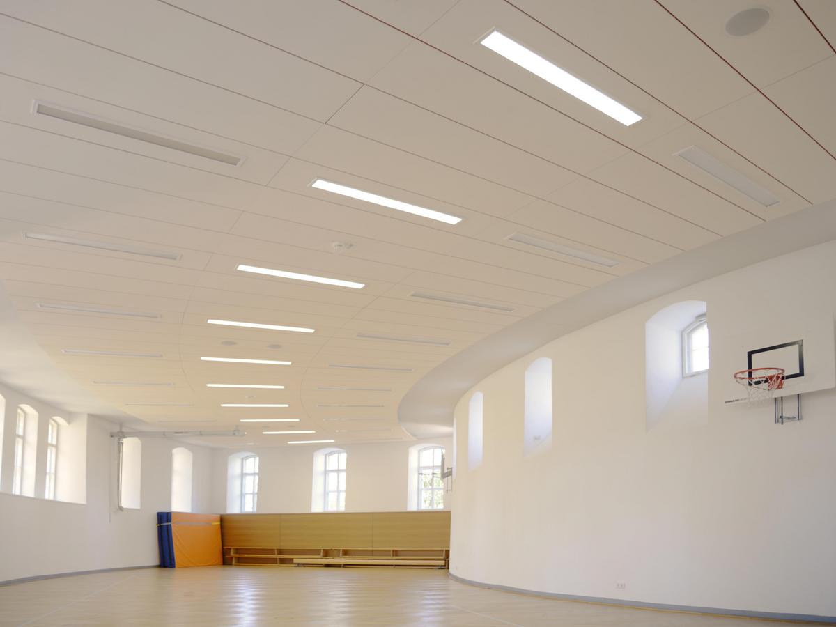Sporthalle mit runden, viereckigen Grundriss. Die Fenster sind weit über Menschenhöhe. Rechts hängt ein Basketballkorb, an der Stirnwand stehen gestapelte Bänke und eine Sprungmatte. Alle Wände weiß, eine Panel-Decke mit langezogenen Lampen.
