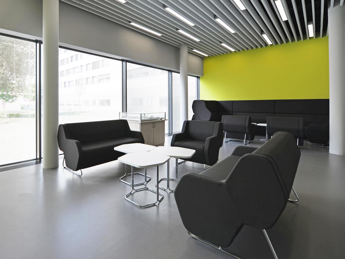 Foyer, großzügiger Raum mit gelber Stirnwand. Großformatigen Fensterverglasungen links, Betonstützen, die dem Raum eine Leichtigkeit geben. Im Raum stehen Sofas und Sessel mit schwarzen Stoff in moderner Formgebung. Der Boden ist dunkelgrau bis schwarz, wahrscheinlich Lino.