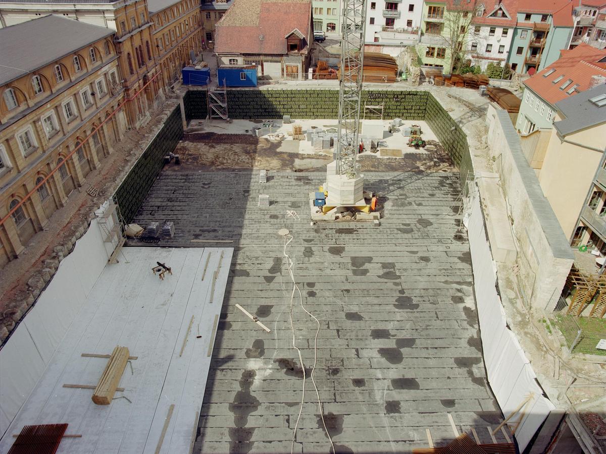 Blick vom Mittelrisalit auf den südlichen Marstallhof. Eine tiefe Baugrube. Ein Kran steht darin und entwächst dem Bild. Die Außenwände sind teilweise mit grüner Dämmung oder weißen Vlies belegt. Die Bodenplatte ist zu zwei Dritteln fertig und mit Bitumen dunkelgrau. Der unsanierte Westflügel rahmt das Bild links.