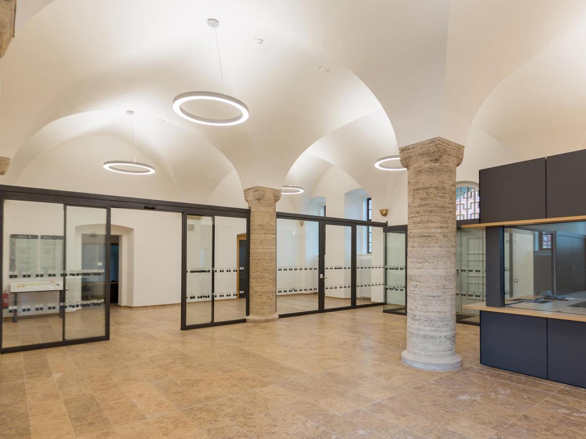 Modernes Foyer mit Rundbogendecken in weiß mit abgehängten kreisrunden Pendelleuchten. Travertin Bodenbelag. Rechts schiebt sich die Wache als dunkelgrauer moderner Kasten in das Bild. Die Stützen sind aus Naturstein und sehr massiv.