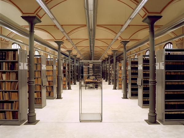 Zentralperspektive mit Stützenreihe links und rechts des Zentrums, die die Rundbögen der Decke aufnehmen. Graue Regale rechts und links davon mit vielen Akten, Ordner und teilweise Büchern alles dunkle gedeckte Farben. Weißer Betonfußboden. Im Zentrum steht eine leere Glasvitrine, dahinter sieht man einen Leseplatz und auch Regale.