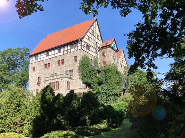 Umgeben von grün und hohen Bäumen und blauen Himmel ist ein hohes Gebäude zu sehen. Die unteren Geschosse sind aus Stein gemauert. Das oberste Geschoss und das Dachgeschoss aus Fachwerk mit weißen Ausfachungen. Rote Ziegeldächer auf allen Gebäudeteilen. Teilweise wachsen die Pflanzen an den Gebäudemauern nach oben.