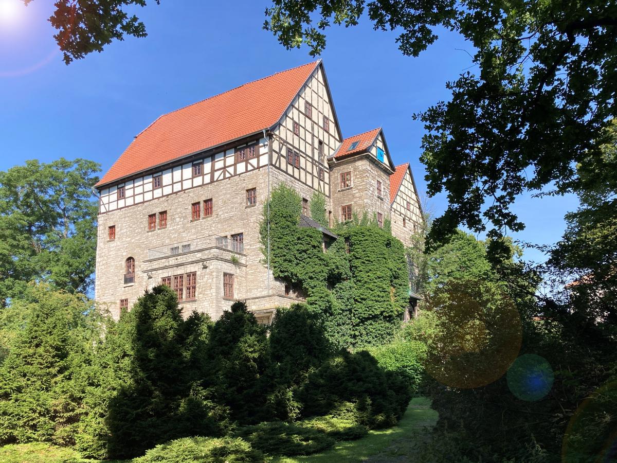 Umgeben von grün und hohen Bäumen und blauen Himmel ist ein hohes Gebäude zu sehen. Die unteren Geschosse sind aus Stein gemauert. Das oberste Geschoss und das Dachgeschoss aus Fachwerk mit weißen Ausfachungen. Rote Ziegeldächer auf allen Gebäudeteilen. Teilweise wachsen die Pflanzen an den Gebäudemauern nach oben.