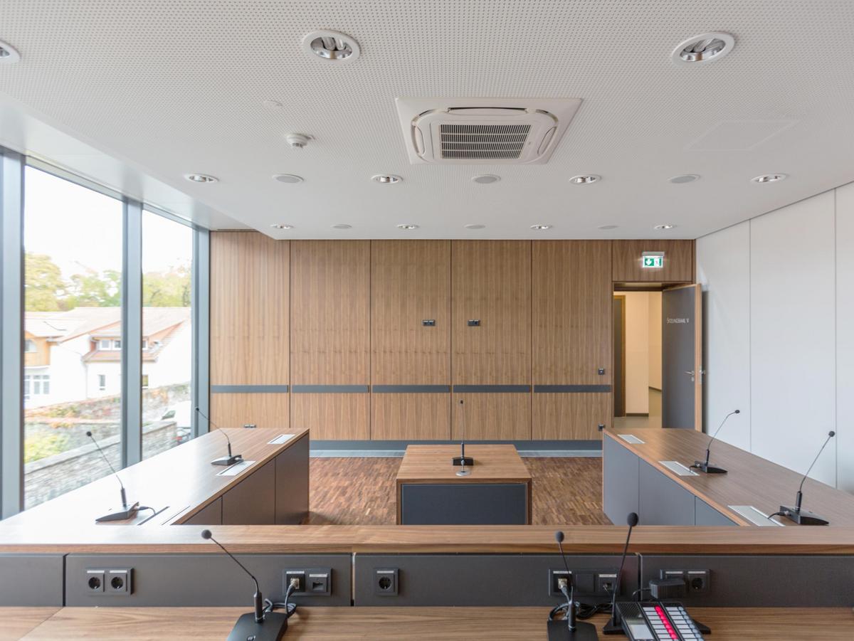 Sitzungssaal, Decke aus Schallschutzplatten gelocht in weiß, links die Glasfassade, vollflächige Verglasung, wir sind im 1. OG. Rechts eine weiße Wand, der Boden aus Stäbchenparkett, dunkel geölt. Im Vordergrund der Richtertisch mit den Mikrofonen. Dahinter die Gegnertische und ein Tisch zur Anhörung in der Mitte.