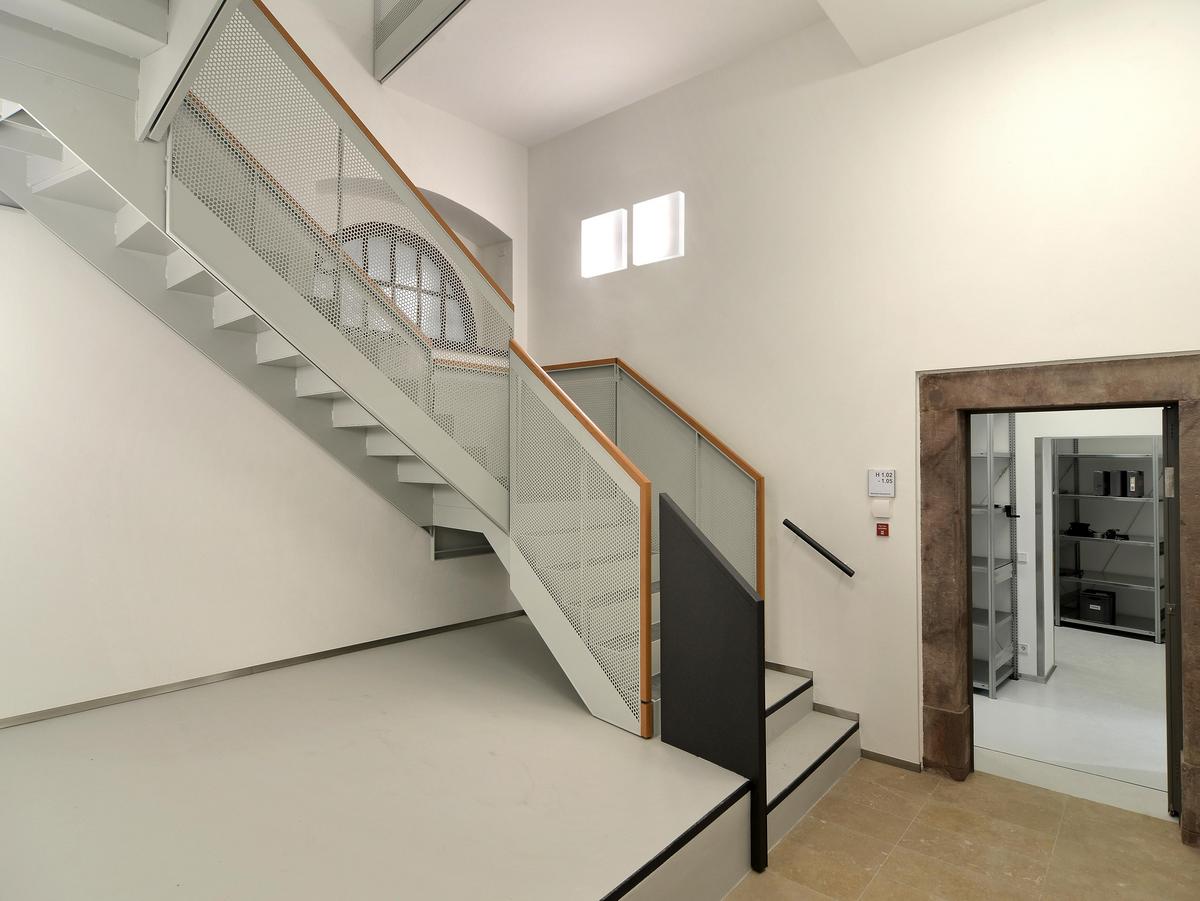 Neues Treppenhaus mit drei Treppenläufen aus Stahl, Wangen und Stufen in lichtgrau. Weiße Wände, rechts geht ein Durchgang in einen Raum mit Regalen, schwere Naturstein-Zarge. Die Treppe hat Brüstungen aus Lochblech mit runder Lochung. Aufgesetzt ist ein Holzhandlauf. Die ersten beiden Stufen haben eine andere Brüstung. Ein schwarze blickdichte Wand als Brüstung die Abstand zur restlichen Brüstung hat.