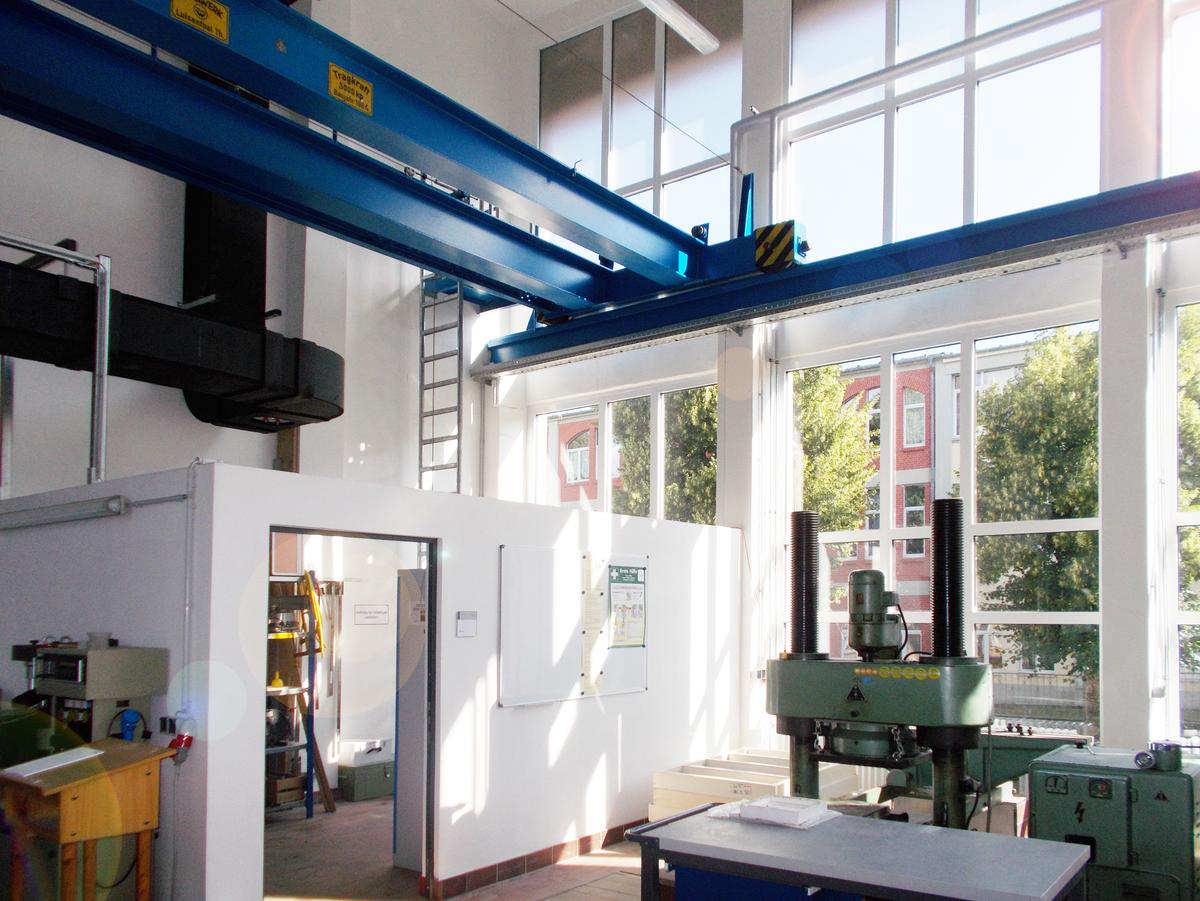 Zweigeschossiger Versuchsraum mit Glasfassade, viel einstrahlendes Sonnenlicht. Schwarze Lüftungsrohre an der Rückwand. Die blaue Krananlage thront über dem Raum und wirkt schwer und dennoch schwebend.