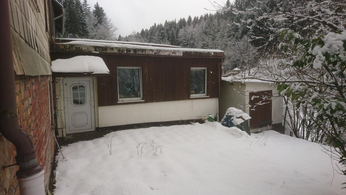 eingeschossiger Anbau mit Holzfassade und Hauseingang; rechts davon leicht hangabwärts eine Garage