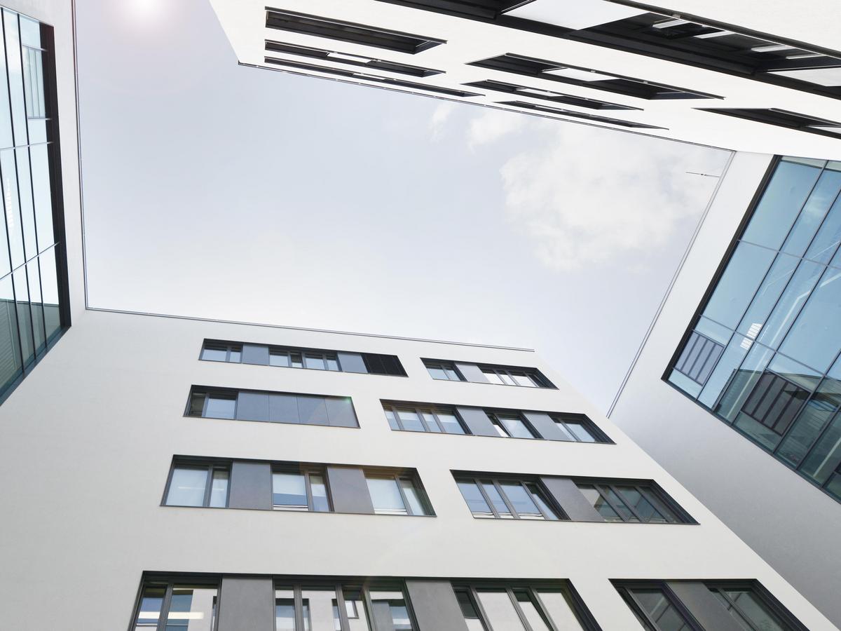 Foto mit Blickrichtung Himmel, leichte Wolken, an allen Bilderrändern Gebäudewände, mindestens vier Geschoss, weiße Putzfassaden mit schwarzen Fensterflächen.