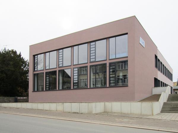 Straßenfassade mit Verglasung ähnlich einer Pfosten-Riegel-Fassade, davor für die Louis-Opel-Straße entlang, rechts kommt der Weg von der Gebäuderückseite an einer kurzen Treppenanlage herunter.