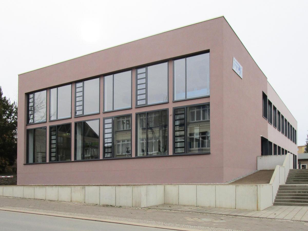 Straßenfassade mit Verglasung ähnlich einer Pfosten-Riegel-Fassade, davor für die Louis-Opel-Straße entlang, rechts kommt der Weg von der Gebäuderückseite an einer kurzen Treppenanlage herunter.