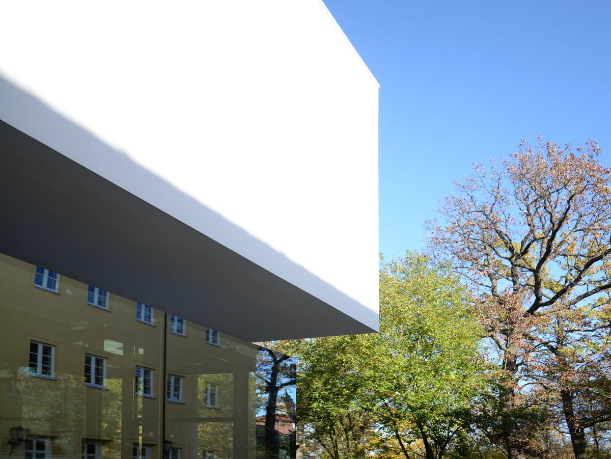 Der weiße Baukörper des Obergeschosses schiebt sich schwer ins Bild und sitz auf der Glasfassade (schwarz) auf und scheint zu schweben. Bei strahlenden Sonnenschein und blauen Himmel sieht man Bäume auf der rechten Bildhälfte.