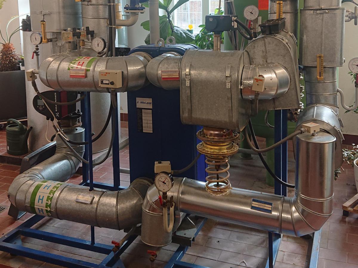 Die Pumpenanlage auf roten Rechteckfliesen, im Hintergrund ein Fenster mit eine Pflanze. Auf einem blauen Gestell ruht die blaue Pumpe in Form eines Kastens von dem sternförmig vier Abgänge sich im Raum verteilen. Die metallischen Rohre haben diverse Aufkleber und Drehräder und Messgeräte.
