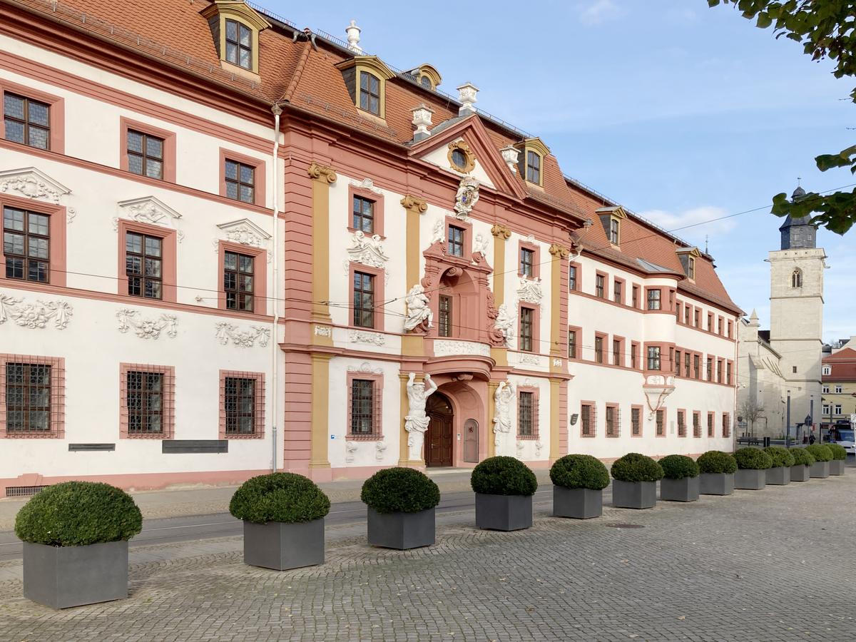 Schräge Ansicht der Patrizierhäuser im Renaissance-Stil. Es dominieren weiße und dunkelrote matte Farben. Alle Faschen, Bänder sind rot gehalten. Die Fassade ist weiß, mit roten Mansarddach mit wenigen Gauben mit gelber Färbung. 3 Geschosse Haupteingang in der Mitte. Davor eine stehen aufgereiht runde Buxbäume in eckigen Töpfen, rechts im Hintergrund ein Kirchturm und eine Straßenbahn.