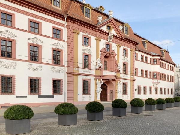Schräge Ansicht der Patrizierhäuser im Renaissance-Stil. Es dominieren weiße und dunkelrote matte Farben. Alle Faschen, Bänder sind rot gehalten. Die Fassade ist weiß, mit roten Mansarddach mit wenigen Gauben mit gelber Färbung. 3 Geschosse Haupteingang in der Mitte. Davor eine stehen aufgereiht runde Buxbäume in eckigen Töpfen, rechts im Hintergrund ein Kirchturm und eine Straßenbahn.