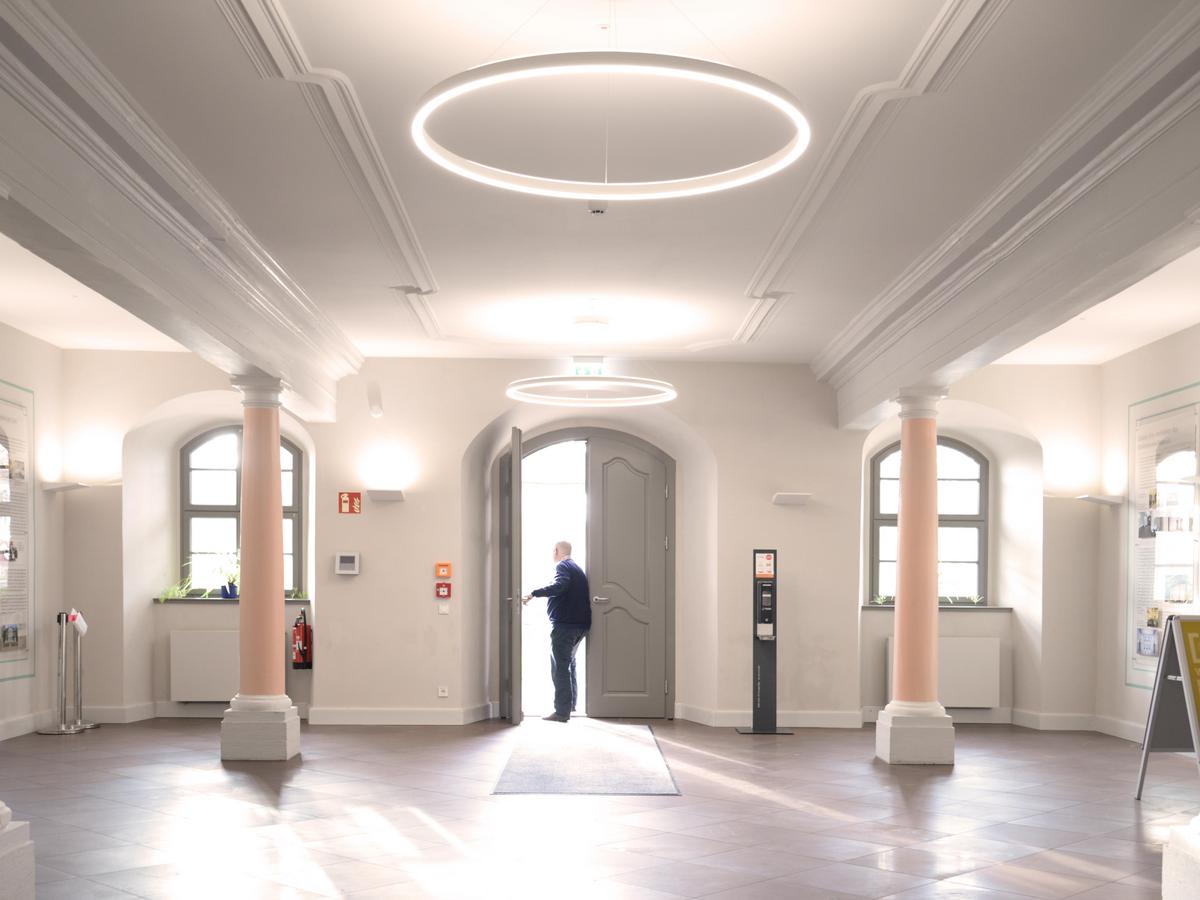 Foyer als Eingang, rechts und links des scheinbares Weges stehen mächtige Säulen in rosafarbenem Farbton, die schwere Deckenbalken tragen, die in weiß, mit Stuck verkleidet sind. Dadurch ergeben sich drei Deckenfelder, die jeweils weiß, mit Stuck verkleidet sind. Die lichtgraue Tür ist zweiflügelig und mit Rundbogen und Kassettierungen. Die beiden Fenster rechts und links davon mit Sprossen und Rundbogen, darunter weiße Heizkörper. Der Fußboden ist mit leicht rötlichen Fliesen belegt.