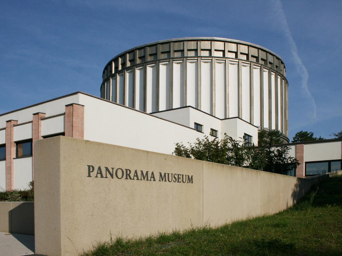 Der Rundbau thront auf den flachen dienen umgebenden Baukörper. Alle in weißer Putzfassade mit Fensteröffnungen. Sehr viele Vor- und Rücksprünge. Im Vordergrund eine Betonwand mit dem Schriftzug des Museums. Rechts davon Grünfläche die nach hinten ansteigt.