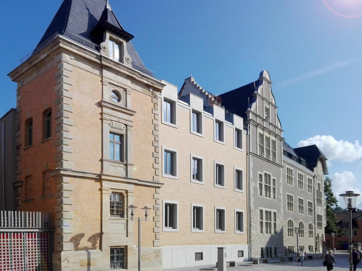 Dreiteilige Gebäudefassade in zarten Orange und Grau mit Risalit und hoch aufragenden Giebel bei strahlend blauen Himmel. Der grau gepflasterte Platz im Vordergrund ist fast menschenleer.