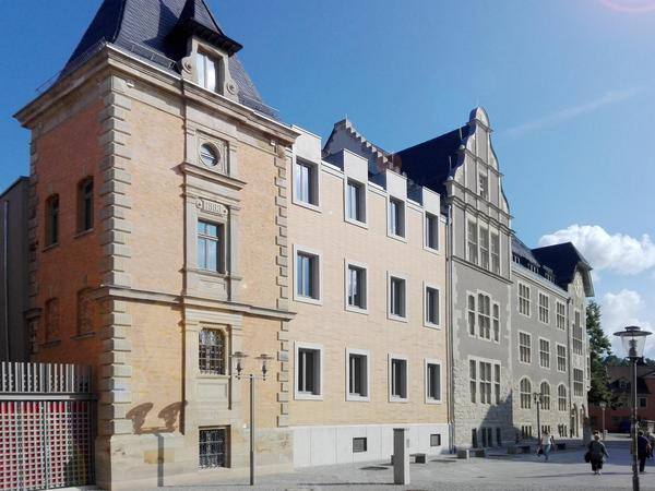 Dreiteilige Gebäudefassade in zarten Orange und Grau mit Risalit und hoch aufragenden Giebel bei strahlend blauen Himmel. Der grau gepflasterte Platz im Vordergrund ist fast menschenleer.