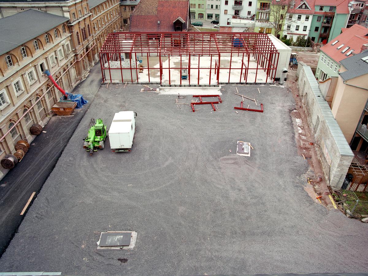Blick vom Mittelrisalit auf den südlichen Marstallhof. Die Baugrube ist geschlossen, eine graue Kiesfläche ist zu sehen. Im oberen Drittel eine Bodenplatte mit roter Stahlskelettstruktur. Hier entsteht der neue Lesesaal im Form der alten Reihalle.