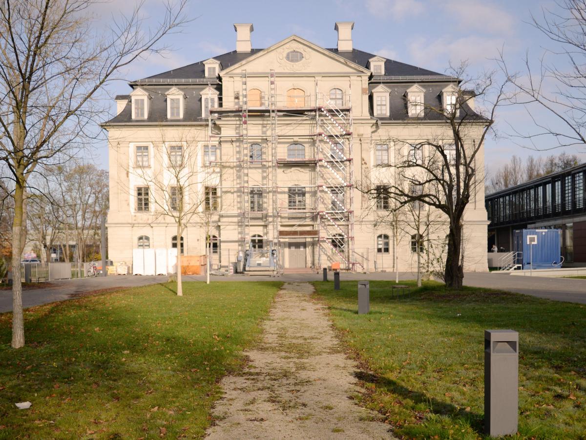 Frontale Südansicht des Barockschlosses. Mittig direkt zulaufender Weg im Vordergrund. Rechts und links davon kahle, herbstliche Bäume, relativ junge Bäume. Klarer, blauer Himmel. Rechts ist der flache Neubau angedeutet sichtbar.