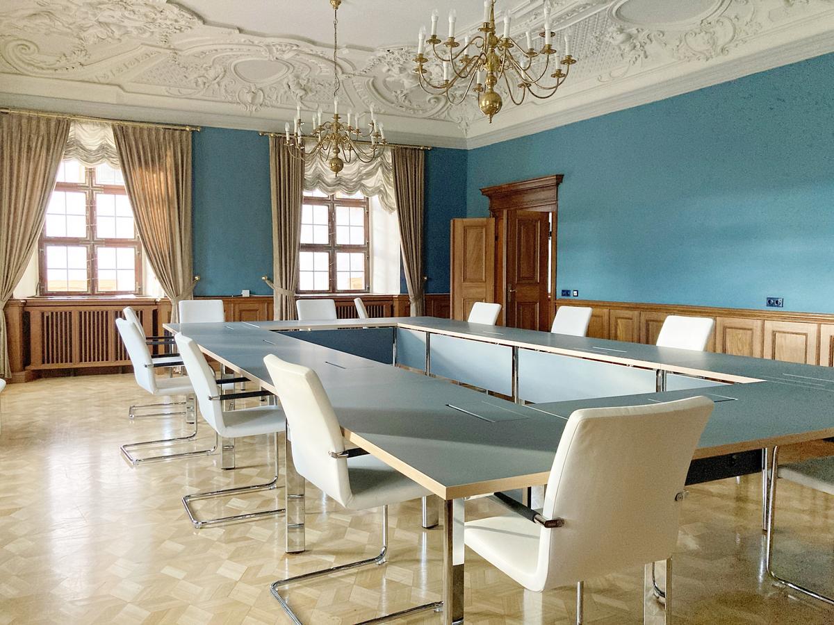Sitzungszimmer mit umlaufenden Lambri aus Holz, darüber naturblaue Farbe, ähnlich Petrol. Die beiden Fenster werden von schweren Vorhängen eingerahmt. Der Boden ist aus Parkett. Im Vordergrund stehen Tische mit ähnlich blauer Tischplatte und weiße Stühle in Null-Aufstellung.