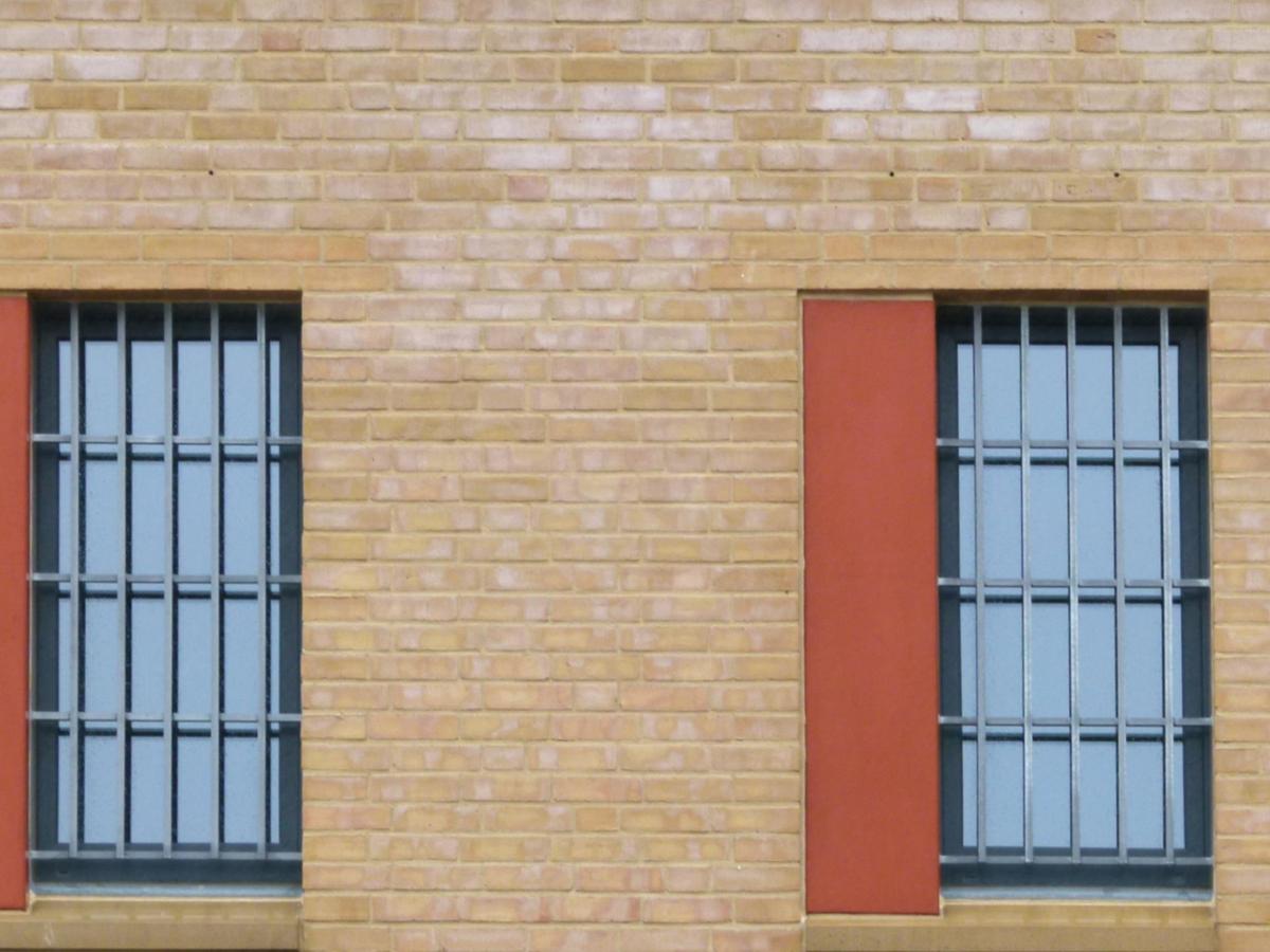 Frontalansicht der Fassade mit zwei Fenster mit grauen Fensterprofilen mit Vergitterung, jeweils links davon ein orangenes Farbfeld in halber Fensterbreite. Sandsteinfarbene Klinkerfassade.