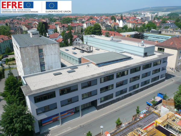 Fassadenansicht von einem gegenüberliegenden Gebäude mit der Straße im Vordergrund von links unten nach rechts mittlere Bildhöhe verlaufend. Das Gebäude ist E-förmig, alle Gebäudeteile mit unterschiedlichen Höhen und Flachdächern. Keine Personen auf der Straße. Im rechten unteren Bilddreieck ist eine Baustelle zu erkennen. Im Hintergrund die Stadt Weimar mit den historischeren Gebäude und rot gedeckten Dächern. In der Ferne das Mahnmal vom KZ Buchenwald.