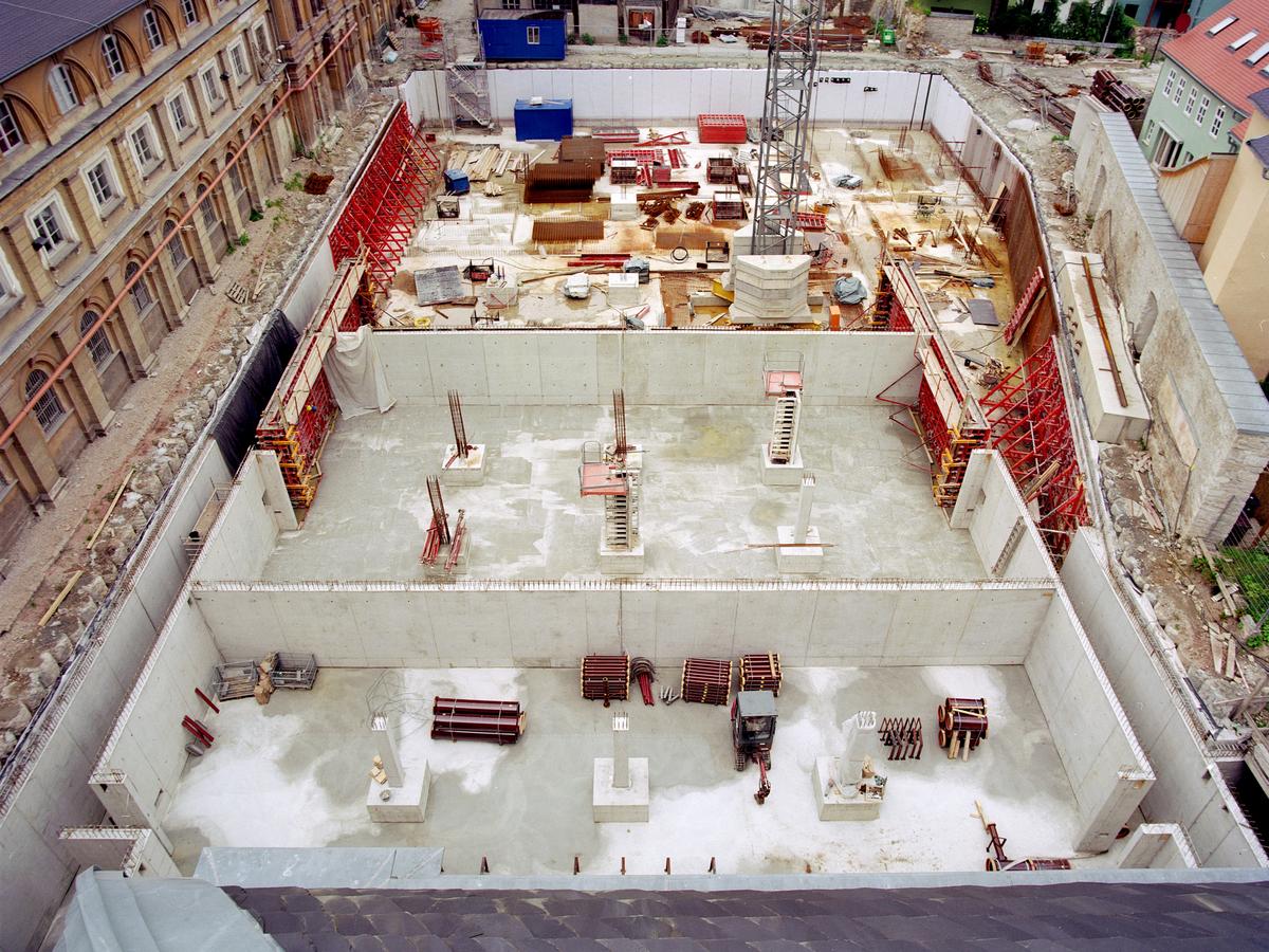 Blick vom Mittelrisalit auf den südlichen Marstallhof. Baugrube ist mit Betonwände eingefasst, die Bodenplatte ist nun in der unteren Hälfte komplett fertig. Teilweise stehen auch schon Beton-Innenwände. Rote Gerüste stützen die Außenwände. Der Westflügel weiter unsaniert Rahmen das Bild links ein.