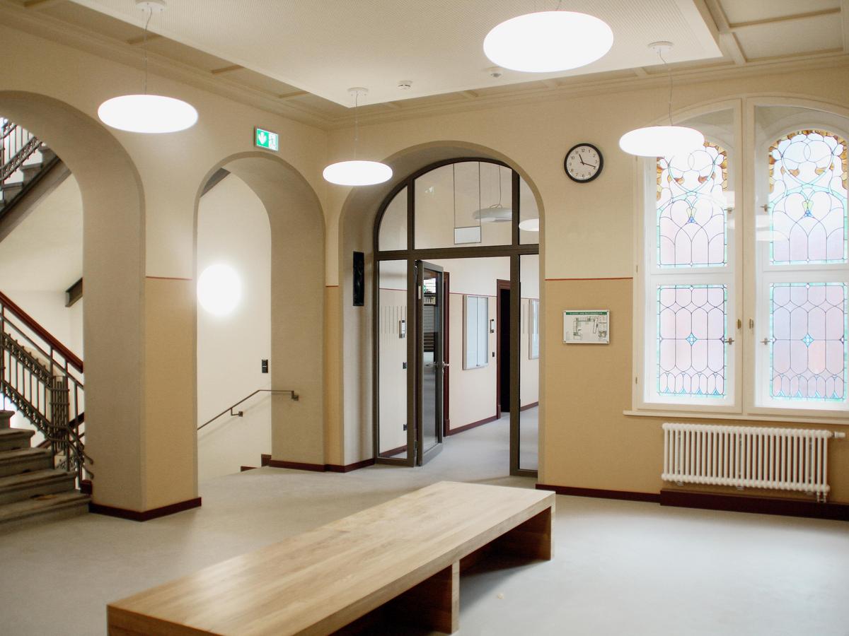 Innenraumfoto mit angeschnittenen Treppenhaus mit schwarzem Jugendstil-Geländer. Die Wände sind hälftig in hellen Brauntönen gemalert. Im Vordergrund eine stilsichere einfache Holz-Sitzbank. Rechts im Bild Bleifenster mit Farbe und Rundbogen, darunter ein weißer Heizkörper, grauer Fußboden.