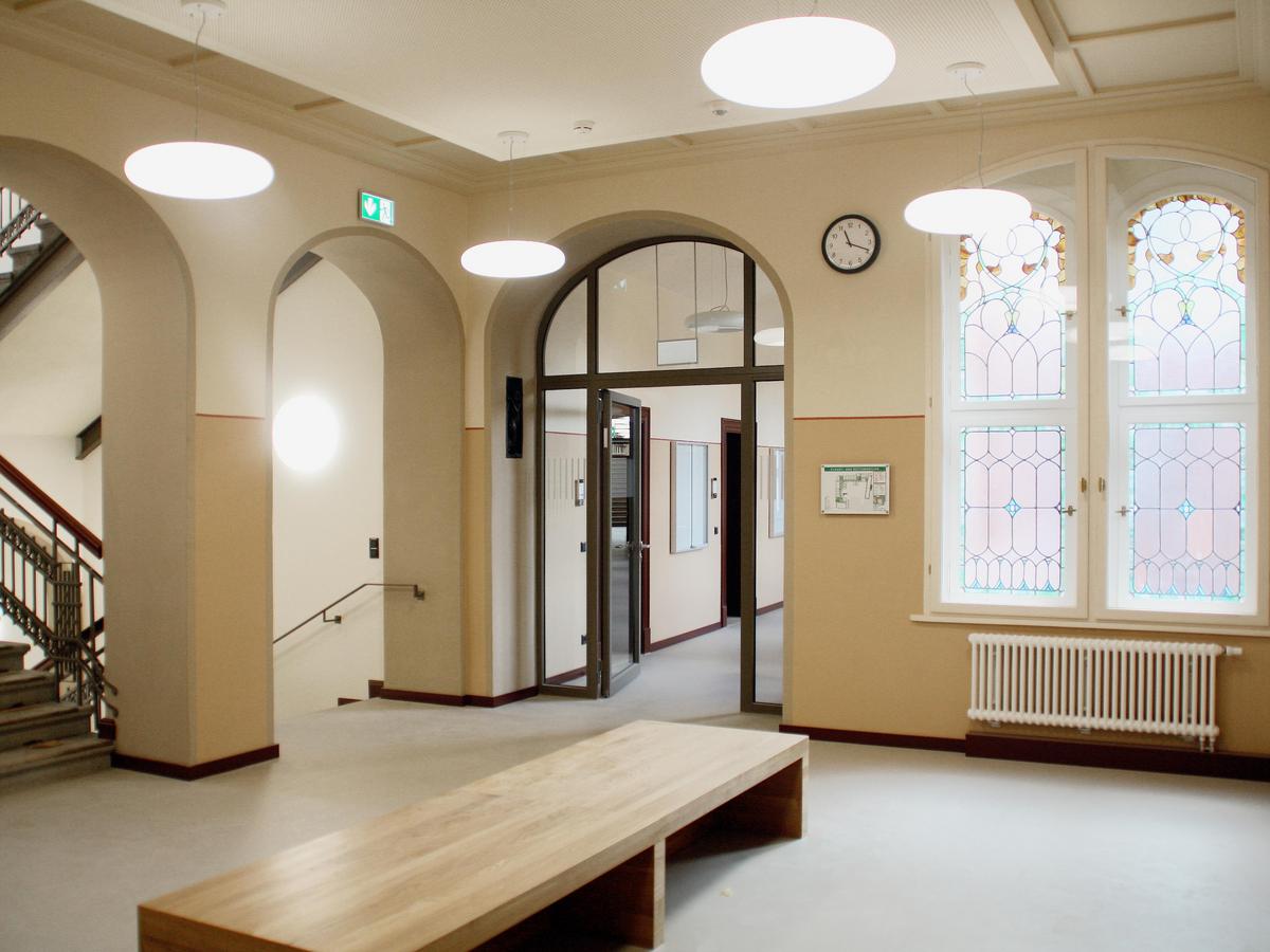 Innenraumfoto mit angeschnittenen Treppenhaus mit schwarzem Jugendstil-Geländer. Die Wände sind hälftig in hellen Brauntönen gemalert. Im Vordergrund eine stilsichere einfache Holz-Sitzbank. Rechts im Bild Bleifenster mit Farbe und Rundbogen, darunter ein weißer Heizkörper, grauer Fußboden.