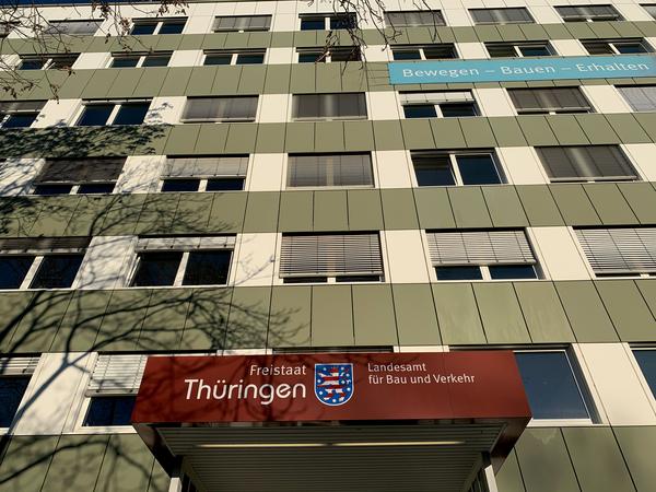 Zu sehen ist der Eingang in das TLBV. Das Gebäude selbst hat eine grün-weiße Fassade mit vielen Fenstern. Über dem Eingangsbereich ist das Wappen des Freistaats Thüringen und die Bezeichnung Freistaat Thüringen, Landesamt für Bau und Verkehr zu lesen.