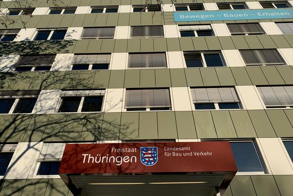Zu sehen ist der Eingang in das TLBV. Das Gebäude selbst hat eine grün-weiße Fassade mit vielen Fenstern. Über dem Eingangsbereich ist das Wappen des Freistaats Thüringen und die Bezeichnung Freistaat Thüringen, Landesamt für Bau und Verkehr zu lesen.