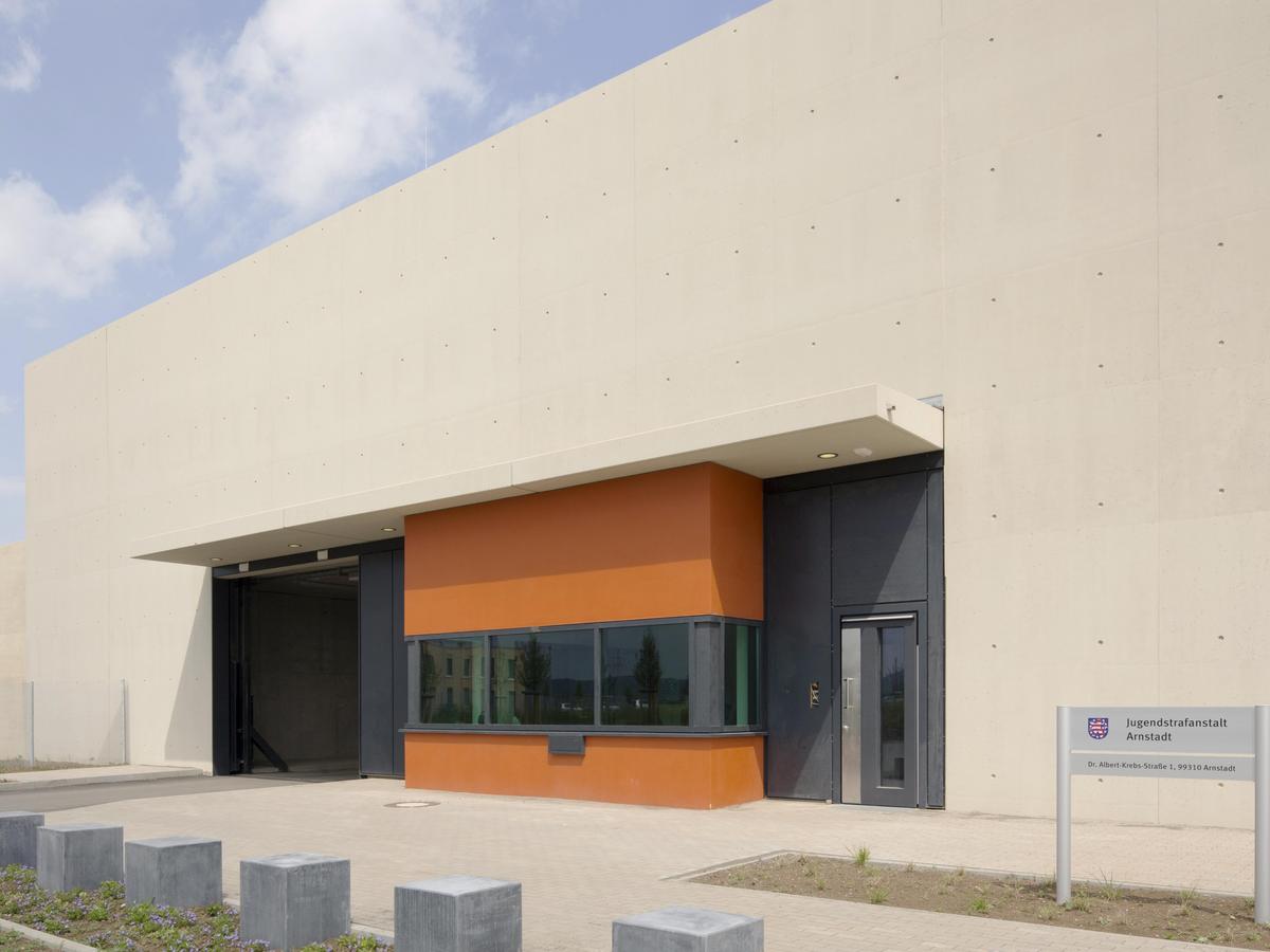 Massives schweres Eingangsgebäude der Strafanstalt mit kurzem Vordach. Geschlossene Fassade, überhöht, sandsteinfarbig mit darauf zuführenden Weg. Orangener Baukörper der Wache schiebt sich in die Fassade. Betonwürfel im Vordergrund.