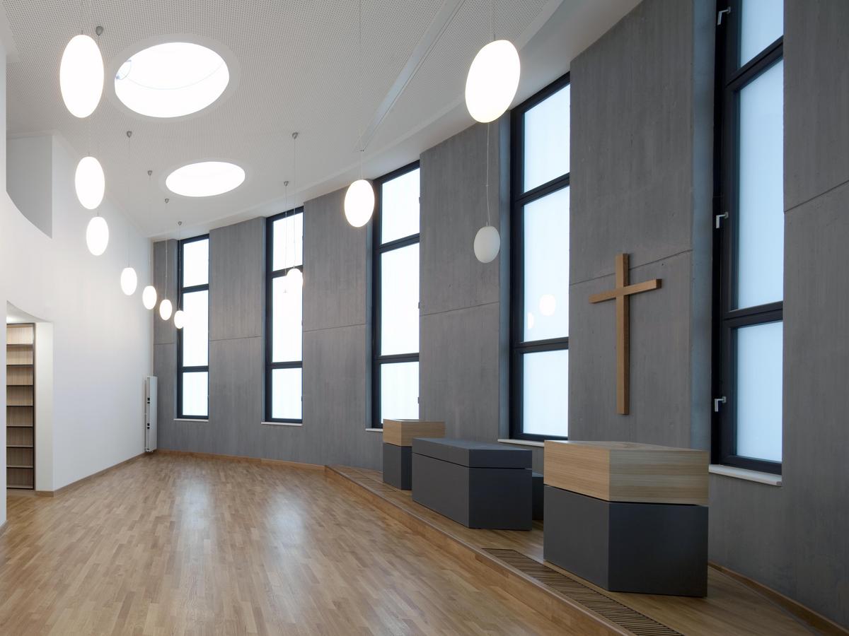 Überhöhter Raum mit augenartigen Grundriss mit Holzparkett, die linke Wand ist weiß mit Zugang in den Raum, die rechte Wand ist grau mit fünf Fenstern und Kreuz an der Wand, davor stehen der Altar und zwei Pulte in grau bzw. Holz. Bohnenartige Lampen hängen von der weißen Decke herab.