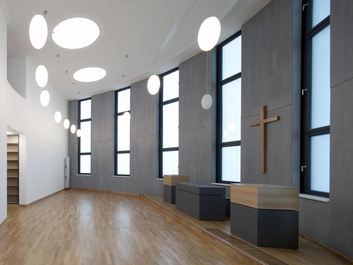 Überhöhter Raum mit augenartigen Grundriss mit Holzparkett, die linke Wand ist weiß mit Zugang in den Raum, die rechte Wand ist grau mit fünf Fenstern und Kreuz an der Wand, davor stehen der Altar und zwei Pulte in grau bzw. Holz. Bohnenartige Lampen hängen von der weißen Decke herab.