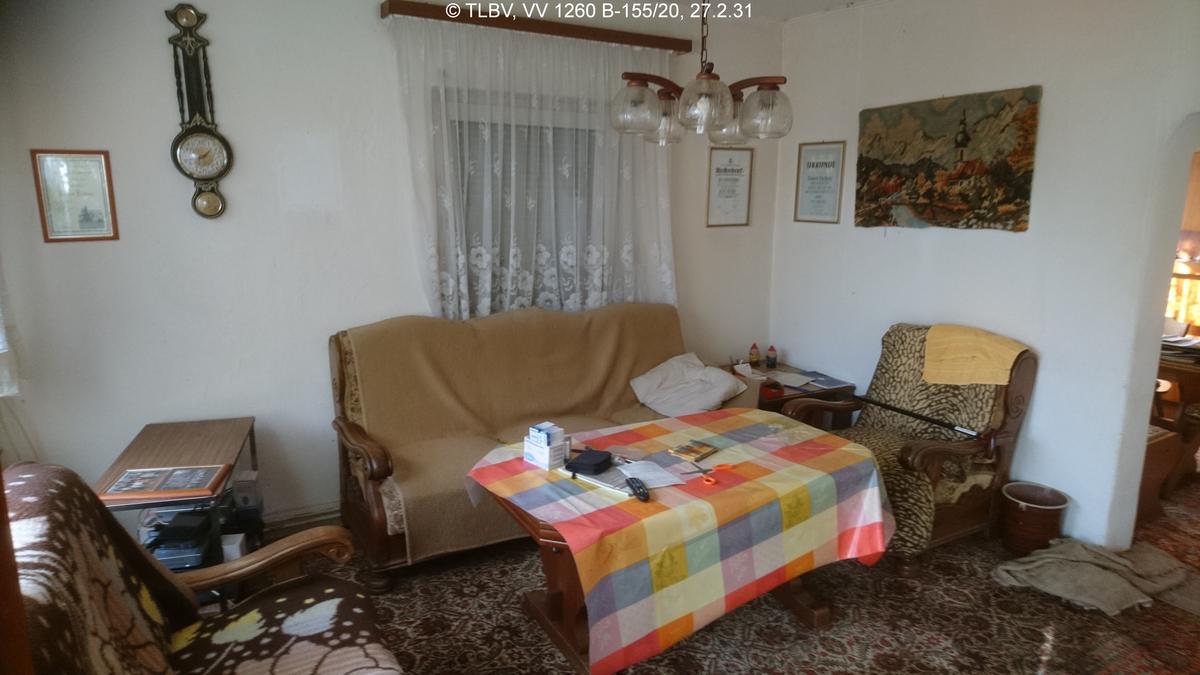 Wohnzimmer, an zwei Seiten Fenster, eins mit heruntergelassener Jalousie, unberäumt