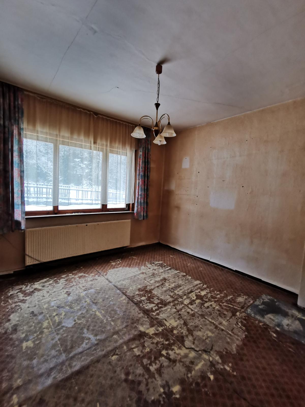Zimmer mit gewelltem Fußbodenbelag, Tapete löst sich am unteren Rand von der Wand ab, ein Fenster, ein Heizkörper
